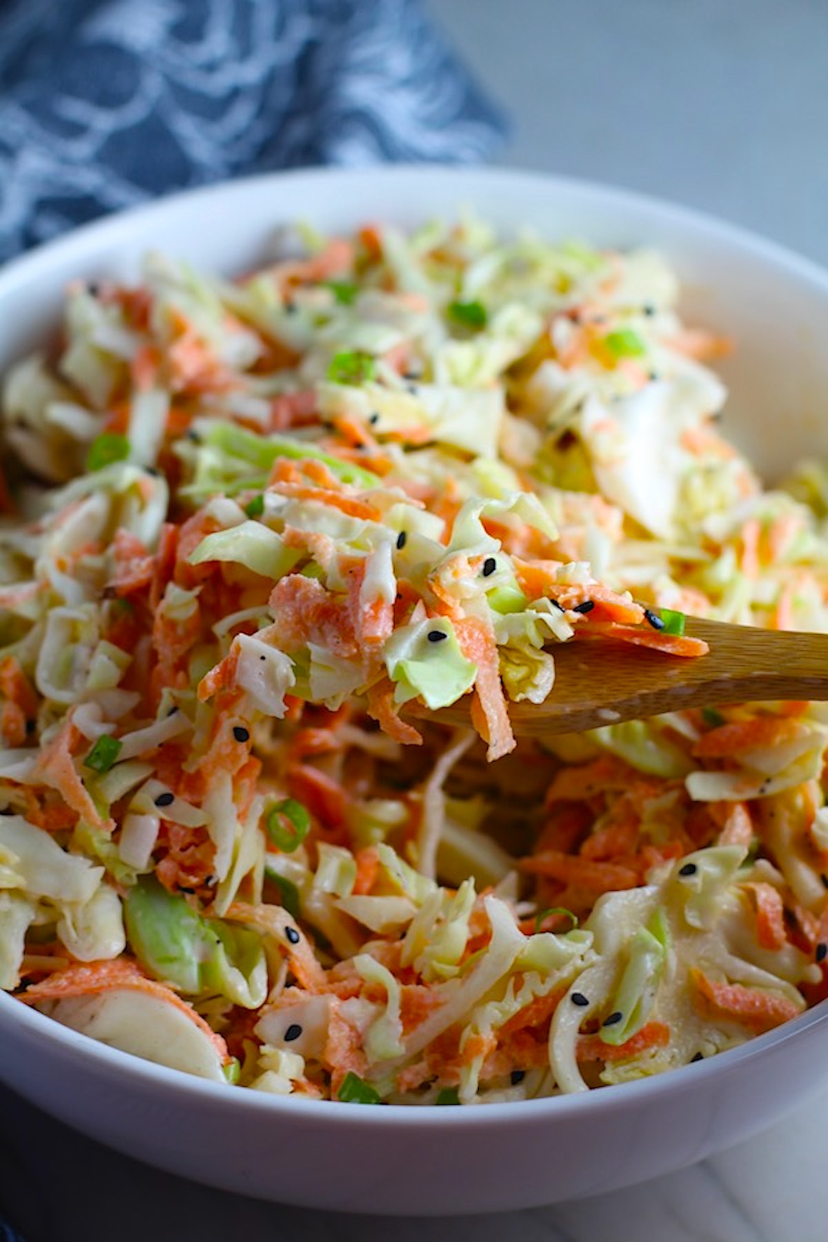 Spoon scooping Coleslaw with an asian salad dressing recipe from a bowl! It has cabbage, carrots, scallions, and sesame seeds in a creamy dressing with mayonnaise. vinegar, soy sauce, and Sesame Oil.  It really is a sidekick that can go with anything!