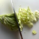Knife slicing cabbage thin for Asian Coleslaw. It has cabbage, carrots, scallions, and sesame seeds in a creamy dressing with mayonnaise. vinegar, soy sauce, and Sesame Oil.  It really is a sidekick that can go with anything!