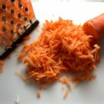 Shredding carrot on a grater for Asian Coleslaw. It has cabbage, carrots, scallions, and sesame seeds in a creamy dressing with mayonnaise. vinegar, soy sauce, and Sesame Oil.  It really is a sidekick that can go with anything!