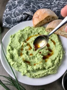 Spoon drizzling Harissa oil over Edamame Dip with Gorgonzola spread on a plate.  It's creamy, savory, nutty, and utterly delicious! 
