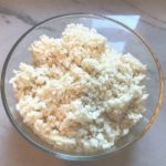Raw Cauliflower Rice in a clear glass bowl on counter for Cheesy Cauliflower Rice recipe.