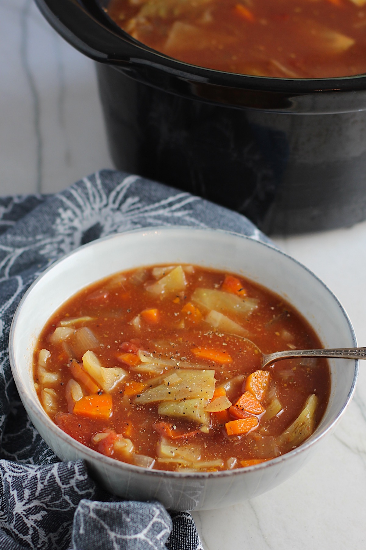 Detox Cabbage Soup recipe in a bowl with spoon on blue napkin on counter with slow cooker in background. The soup is tomato-based with chunky veggies: cabbage, tomato, carrots, onion, and garlic.