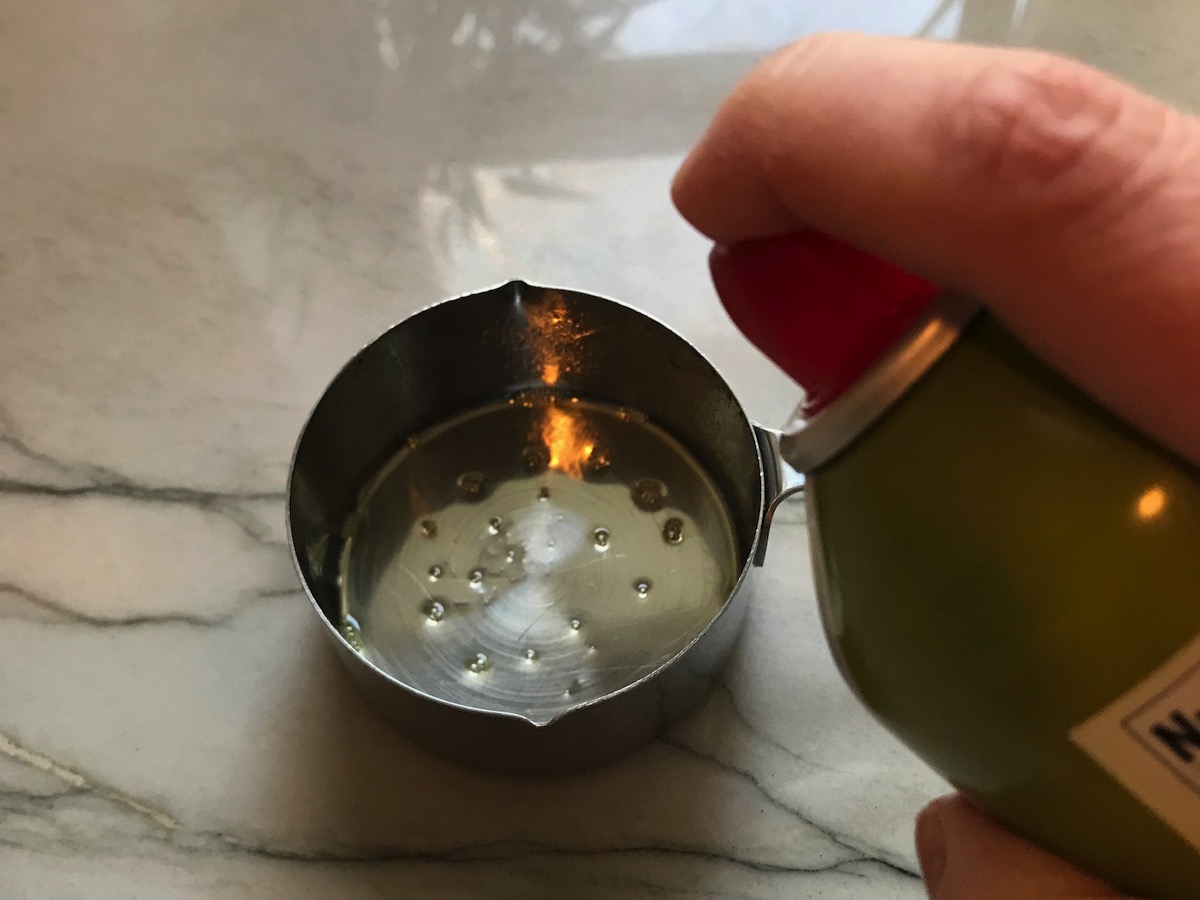 Hand spraying olive oil into a measuring cup for Best Honey Ketchup recipe.