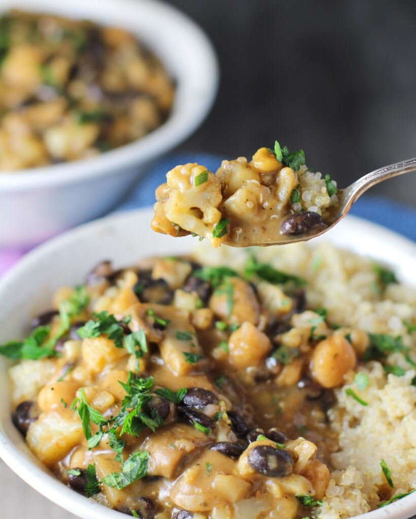 Spoonful of Creamy Cajun Cauliflower and Beans over Quinoa over a filled bowl. The mild cajun flavors combined with  healthy garbanzo beans, black beans, cauliflower, and mushrooms make this a hearty, creamy, and delicious dish. 