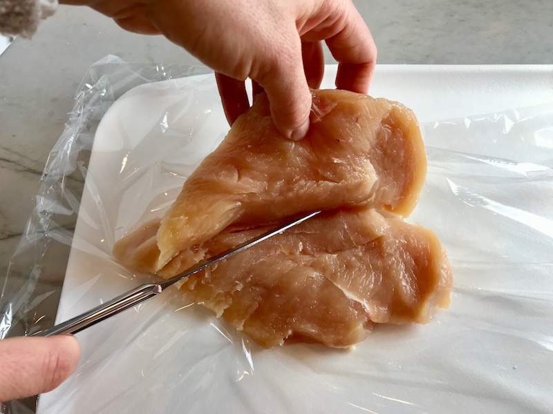 Hands holding and slicing a chicken breast from the side to butterfly it.