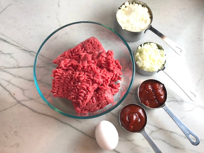 Raw Ground Beef in bowl with parmesan cheese, onions and ketchup in measuring cups on counter and egg on counter. This 5-Ingredient Easy Meatloaf recipe may be short on ingredients, but it is loaded with flavor. The lack of breadcrumbs means you don't have them soaking up the juices, so the meat stays moist - and it's gluten free too! Parmesan cheese, onion, egg, ketchup, salt, and pepper are all that go into this yummy loaf of goodness!
