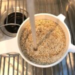 Uncooked white quinoa in a strainer with running water rinsing it in over sink for Creamy Parmesan Mushroom Quinoa Risotto.