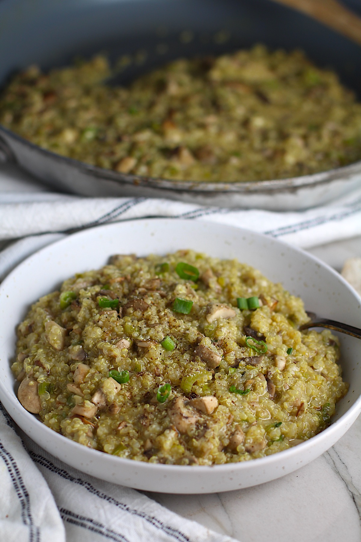 Creamy Parmesan Mushroom Quinoa Risotto in a bowl with fork and skillet in background. This recipe has the traditional garlic flavor in the background and the nuttiness from the  Parmesan combined with the meaty earthy flavor of the mushrooms.  There is no wine.