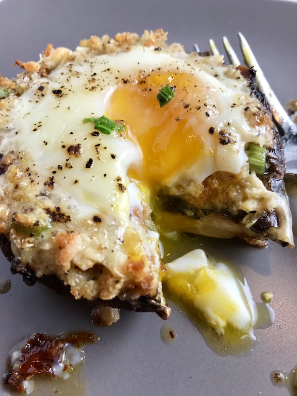 Portabella Mushroom Egg Nests on plate with bite cut out and runny yoke and fork on plate. Portabella mushrooms are filled with garlic, scallion, breadcrumb & Manchego cheese stuffing!  Then an egg is baked nestled in the center of all of this goodness.