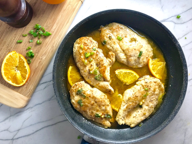 Orange Brown Butter Sauce Chicken with orange slices and parsley garnish on top in skillet on counter with wood cutting board in background with sliced oranges and parsley.