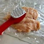 Hand using meat tenderizer to pound out chicken breast.