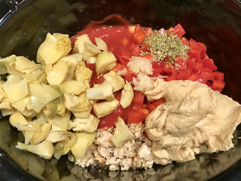 Artichoke pieces, diced tomatoes, seasoning, hummus, and cooked ground chicken in a slow cooker insert before cooking for Chicken ragu recipe.