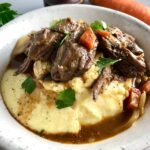 Cooked short ribs on top of creamy polenta in a bowl with sauce, carrots, and onions. Bowl sitting on counter.