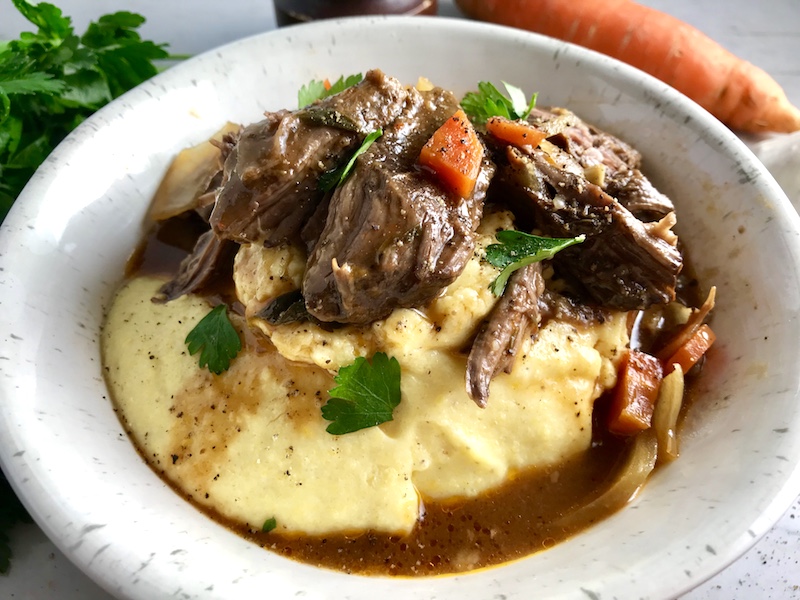 Cooked short ribs on top of creamy polenta in a bowl with sauce, carrots, and onions. Bowl sitting on counter.