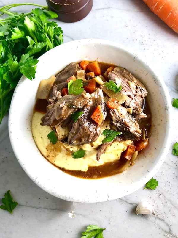 Cooked short ribs on top of creamy polenta in a bowl with sauce, carrots, and onions for flanken short ribs recipe slow cooker. Bowl sitting on counter.