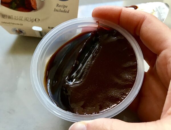 Hand holding dark brown colored demi glace in a plastic cup for flanken short ribs recipe slow cooker.