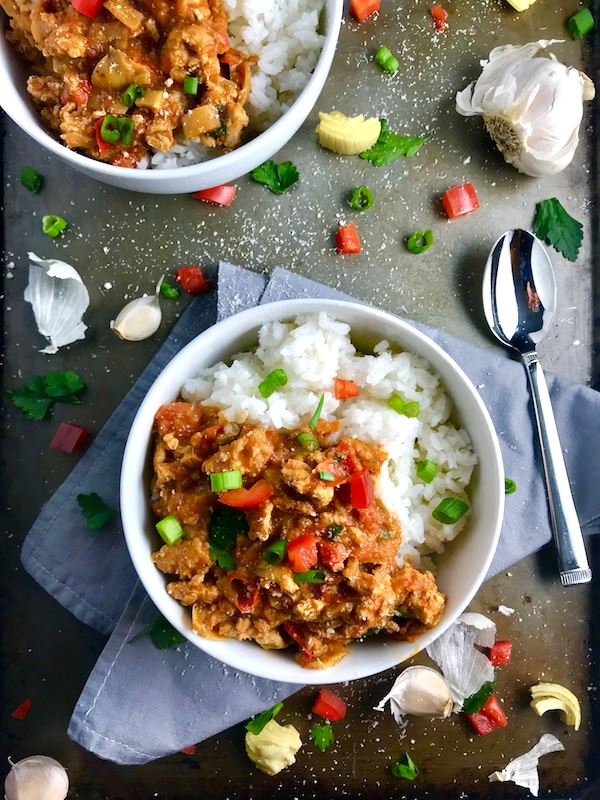 Slow cooker Ground chicken ragu in a bowl over rice with diced red pepper, scallions, and pieces of artichoke and garlic cloves on counter with a spoon.
