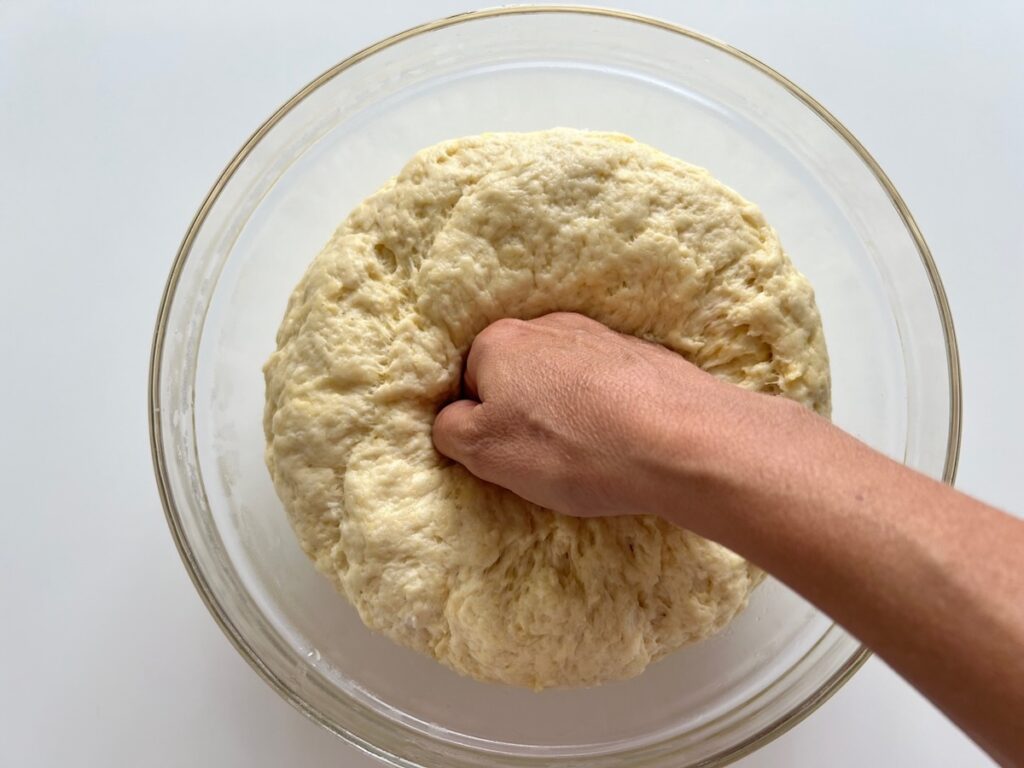 Hand 'punching' down the risen dough in a large glass bowl so it can rise for Chicago Pizza with Chicken Sausage.