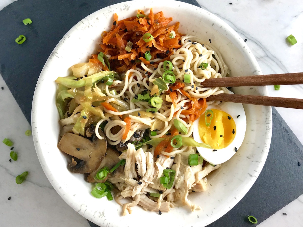 Healthy Chicken Ramen Noodles  in a bowl with chopsticks and Chicken, carrots, cabbage, half of a hard boiled egg, scallions, and black sesame seeds.