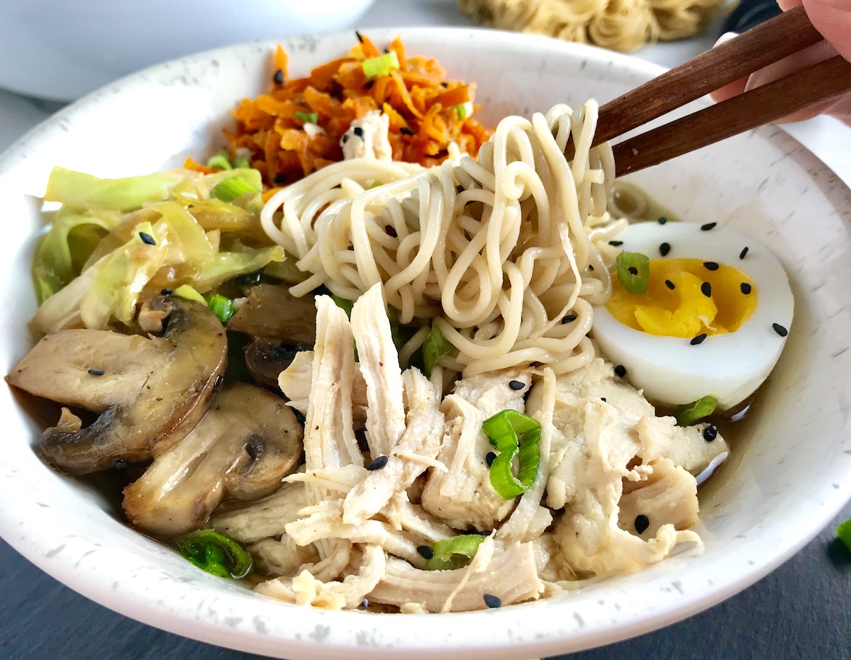 Healthy Chicken Ramen Noodles in a bowl with chopsticks.  The bowl has Chicken, carrots, cabbage, half of a hard boiled egg, scallions, and black sesame seeds.