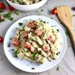Noodles with cauliflower cream sauce, sausage, and tomatoes on white plate on counter with tomatoes, parsley, and spoon on counter