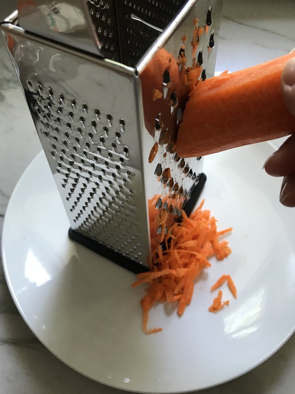 Whole carrot being shredded on a grater for Healthy Chicken Ramen bowls.
