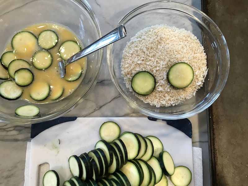 Dipping raw zucchini into egg and into breadcrumbs.
