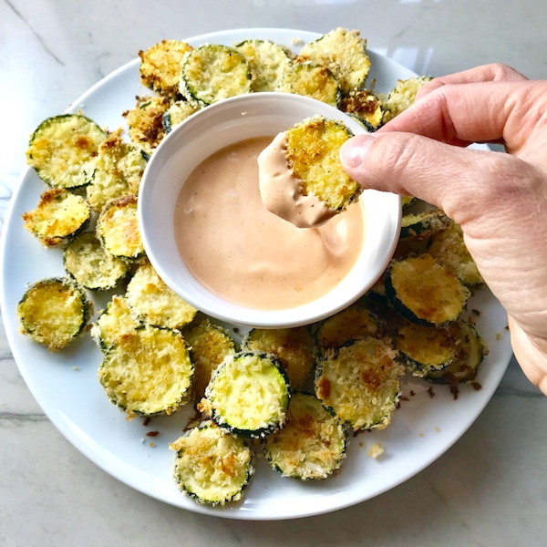 hand holding zucchini chip after dipping in sauce. Plate with chips and dip.  Learn how to roast zucchini slices.