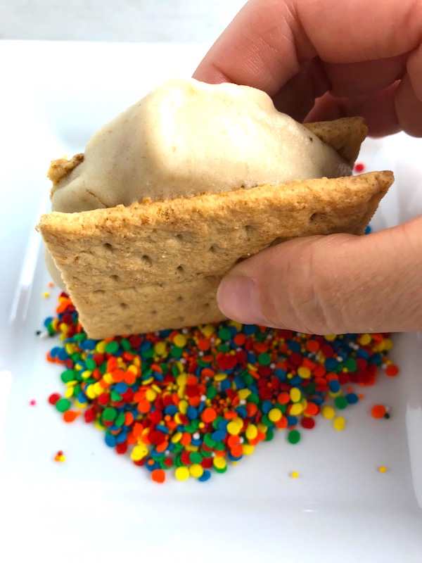 Dipping a banana and graham cracker ice cream sandwich into rainbow sprinkles. Banana Ice Cream is literally just 2 ingredients, Frozen Bananas and Almond Milk.  Add in crunchy Graham Crackers as the 3rd ingredient and you have a delicious healthy Banana Ice Cream Sandwich!  If you have never tried banana ice cream, you are in for a treat!  It tastes just like soft serve ice cream.