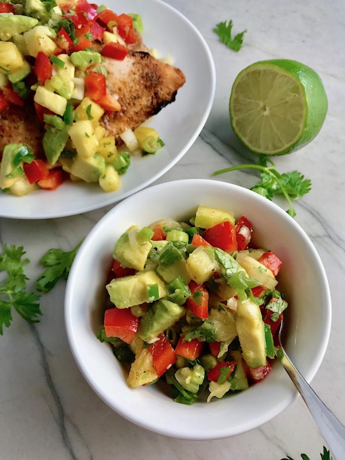 Pineapple Salsa with red pepper, jalapeño, cilantro, scallion, lime, and avocado in a bowl with a spoon on the counter with a plate of grilled chicken and salsa in back and half of a lime on counter.