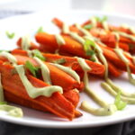 Close up of roasted carrots on plate with avocado crema drizzled on top with scallion slices. Roasted Carrots with smoky cumin and scallions, then topped with Avocado Crema are such a delicious and quick side dish for dinner.  Roasting brings out the natural sugar in the carrots, so they get a sweet caramelization with the salty and smoky flavors. The Avocado crema gives creamy and silky balance. 