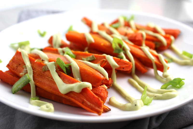 Close up of roasted carrots on plate with avocado crema drizzled on top with scallion slices. Roasted Carrots with smoky cumin and scallions, then topped with Avocado Crema are such a delicious and quick side dish for dinner.  Roasting brings out the natural sugar in the carrots, so they get a sweet caramelization with the salty and smoky flavors. The Avocado crema gives creamy and silky balance. 