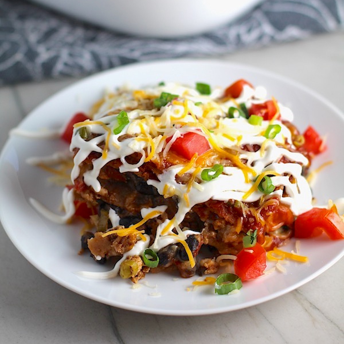 Piece of Ground Chicken taco casserole on plate with sour cream drizzled on top and diced tomatoes and scallion on top. What could be better than tacos? A Taco Casserole that's made with healthier lean ground chicken and can be made ahead for your busy schedule. This Taco Casserole has corn tortillas layered with browned ground chicken seasoned with smokey mexican spices, black beans, tomatoes, and cheese, and then more cheese.