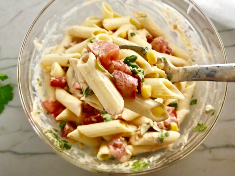 Clear bowl on counter with mixed Mexican Pasta Salad with corn, tomatoes, pasta, Mexican dressing, and cilantro. This Creamy Mexican Pasta Salad has Sun Dried Tomatoes, Corn, Fresh Diced Tomatoes, Cilantro, and smoky Mexican spices. The dressing is creamy and smoky with lots of depth and a hint of sweetness from the Sun Dried Tomatoes that are blended in. Terrific as a side or mix in some chicken or beef and call it a meal!