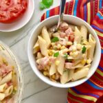 Close up of Mexican Pasta Salad in white bowl on counter with second bowl in upper left corner and tomato half on plate. This Creamy Mexican Pasta Salad has Sun Dried Tomatoes, Corn, Fresh Diced Tomatoes, Cilantro, and smoky Mexican spices. The dressing is creamy and smoky with lots of depth and a hint of sweetness from the Sun Dried Tomatoes that are blended in. Terrific as a side or mix in some chicken or beef and call it a meal!