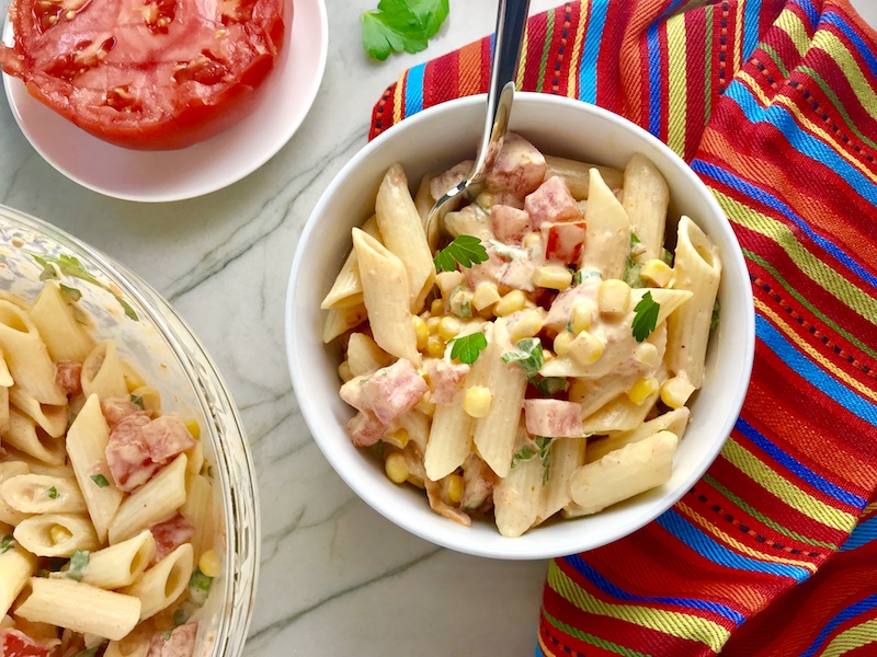 Close up of Mexican Pasta Salad in white bowl on counter with second bowl in upper left corner and tomato half on plate. This Creamy Mexican Pasta Salad has Sun Dried Tomatoes, Corn, Fresh Diced Tomatoes, Cilantro, and smoky Mexican spices. The dressing is creamy and smoky with lots of depth and a hint of sweetness from the Sun Dried Tomatoes that are blended in. Terrific as a side or mix in some chicken or beef and call it a meal!