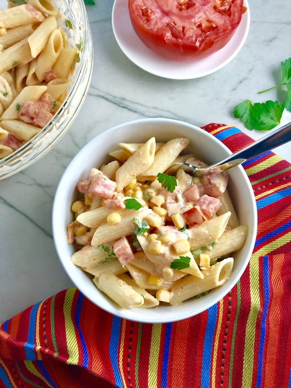 Close up of Mexican Pasta Salad in white bowl on counter with second bowl in upper left corner and tomato half on plate. This Creamy Mexican Pasta Salad has Sun Dried Tomatoes, Corn, Fresh Diced Tomatoes, Cilantro, and smoky Mexican spices. The dressing is creamy and smoky with lots of depth and a hint of sweetness from the Sun Dried Tomatoes that are blended in. Terrific as a side or mix in some chicken or beef and call it a meal!