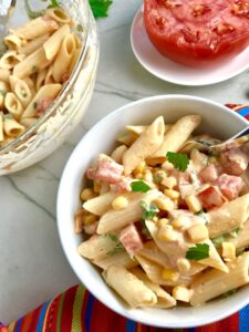 Close up of Mexican Pasta Salad in white bowl on counter with second bowl in upper left corner and tomato half on plate. This Creamy Mexican Pasta Salad has Sun Dried Tomatoes, Corn, Fresh Diced Tomatoes, Cilantro, and smoky Mexican spices. The dressing is creamy and smoky with lots of depth and a hint of sweetness from the Sun Dried Tomatoes that are blended in. Terrific as a side or mix in some chicken or beef and call it a meal!