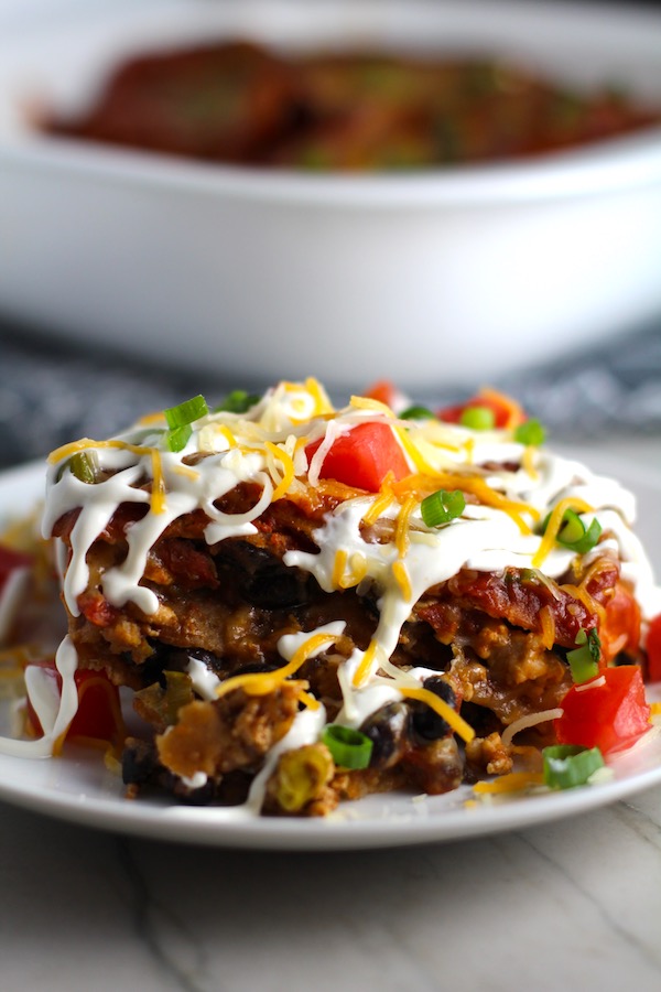 Close up side view of taco casserole piece on plate with sour cream drizzled on top and diced tomatoes and scallion on top. What could be better than tacos? A Taco Casserole that's made with healthier lean ground chicken and can be made ahead for your busy schedule. This Taco Casserole has corn tortillas layered with browned ground chicken seasoned with smokey mexican spices, black beans, tomatoes, and cheese, and then more cheese.