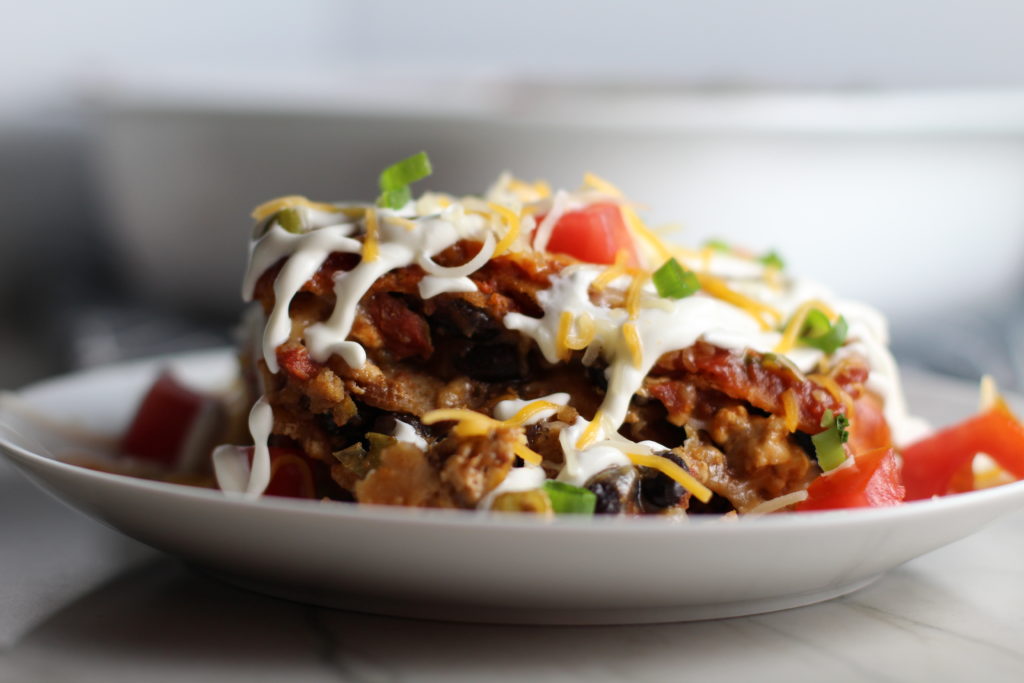 Close up side view of taco casserole piece on plate with sour cream drizzled on top and diced tomatoes and scallion on top. What could be better than tacos? A Taco Casserole that's made with healthier lean ground chicken and can be made ahead for your busy schedule. This Taco Casserole has corn tortillas layered with browned ground chicken seasoned with smokey mexican spices, black beans, tomatoes, and cheese, and then more cheese.