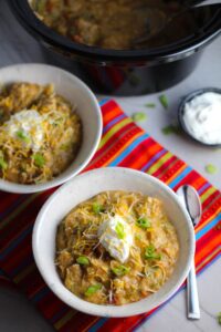 2 Bowls of Chipotle Chicken Enchilada Stew with sour cream and scallion slices as garnish and slow cooker pot in background. It's one of my Prepped Freezer Meal recipes and it's creamy, cozy, hearty, cheesy, and slightly spicy. All ingredients freeze raw, thaw, then cook in the slow cooker or in the oven. It's a stew because it's thick and hearty with bites of chicken, carrots, onion, chipotle peppers, cumin, garlic and tortilla chips that melt down to thicken this delicious stew.