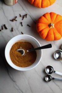 Pumpkin Spice blend in white bowl with silver spoon on counter with mini pumpkins, cloves, and measuring spoons. This Pumpkin Spice Recipe has 4 simple ingredients and takes minutes to make! PLUS, here are 20 EASY WAYS TO USE this Spice blend, many with no baking at all!