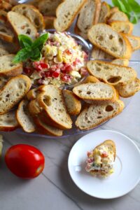 Bruschetta Topping surrounded by crostini on a platter with one crostini topped on a plate. It combines fresh tomatoes, artichoke hearts, garlic, basil, olive oil, and GOAT CHEESE!Â  Top on toasty garlic crostini and you get fresh bright flavors with both crunch and creamy in every bite.