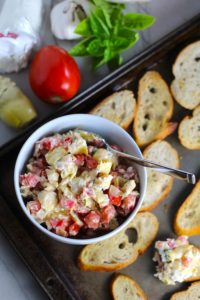 Bruschetta Topping in bowl on pan surrounded by crostini with ingredients to the left. It combines fresh tomatoes, artichoke hearts, garlic, basil, olive oil, and GOAT CHEESE!  Top on toasty garlic crostini and you get fresh bright flavors with both crunch and creamy in every bite.