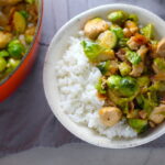 Easy Bacon Brussel Sprouts and Chicken served over white rice in a bowl with orange pot in background. It is a perfect quick Fall Recipe!  It has a creamy sauce filled with salty bacon, earthy and almost nutty seared brussel sprouts, and hearty healthy chicken. Serve over rice (or Quinoa or pasta!).