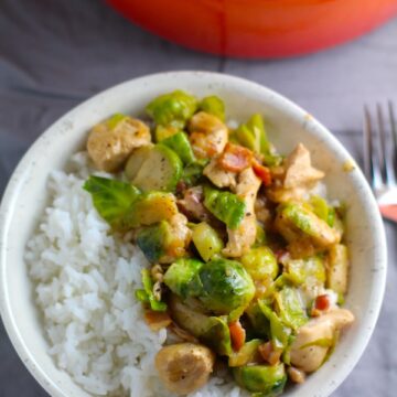 Chicken Brussel Sprouts Bacon served over white rice in a bowl with orange pot in background. It is a perfect quick Fall Recipe!  It has a creamy sauce filled with salty bacon, earthy and almost nutty seared brussel sprouts, and hearty healthy chicken. Serve over rice (or Quinoa or pasta!).