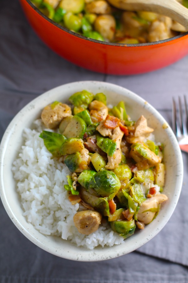 Chicken, Brussels Sprouts, and Bacon served over white rice in a bowl with orange pot in background. It is a perfect quick Fall Recipe!  It has a creamy sauce filled with salty bacon, earthy and almost nutty seared brussel sprouts, and hearty healthy chicken. Serve over rice (or Quinoa or pasta!).