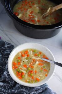 5-Ingredient Homemade Chicken Soup in a bowl with spoon and crockpot in background. This soup is thick, creamy, and hearty. It's filled with flavorful broth, meaty shredded chicken, and sweet bursts of bright veggies. You can prepare all of the ingredients ahead and freeze them until you are ready to cook. Then, thaw, pour into the Slow Cooker, set and forget.