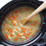 5-Ingredient Homemade Chicken Soup in a crockpot with wood spoon. This soup is thick, creamy, and hearty. It's filled with flavorful broth, meaty shredded chicken, and sweet bursts of bright veggies. You can prepare all of the ingredients ahead and freeze them until you are ready to cook. Then, thaw, pour into the Slow Cooker, set and forget.