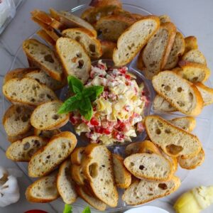 Bruschetta Topping surrounded by crostini on a platter with one crostini topped on a plate. It combines fresh tomatoes, artichoke hearts, garlic, basil, olive oil, and GOAT CHEESE!Ā Top on toasty garlic crostini and you get fresh bright flavors with both crunch and creamy in every bite.